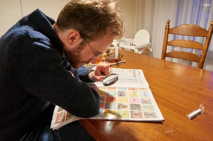 me photographed from the side using a looking glass to look at a puzzle in a newspaper